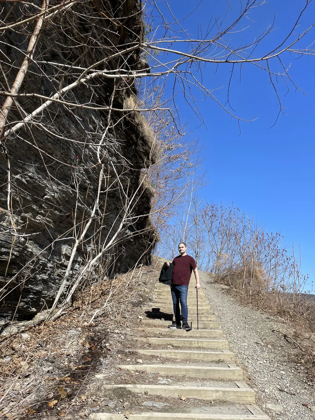 Me standing on a staircase next to a shale cliff. I'm wearing a maroon T-shirt and jeans, carrying a black coat, and using a maroon cane to stand.