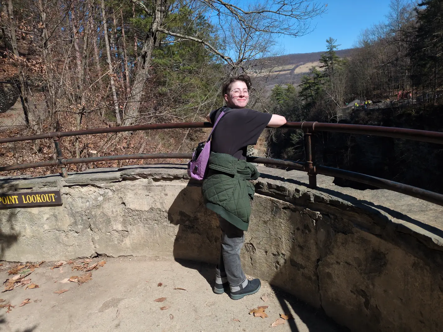 My partner at a railing, smiling back at the camera. They're wearing wire-rimmed glasses, black T-shirt and jeans, a small purple backpack, and a green coat tied around their waist.