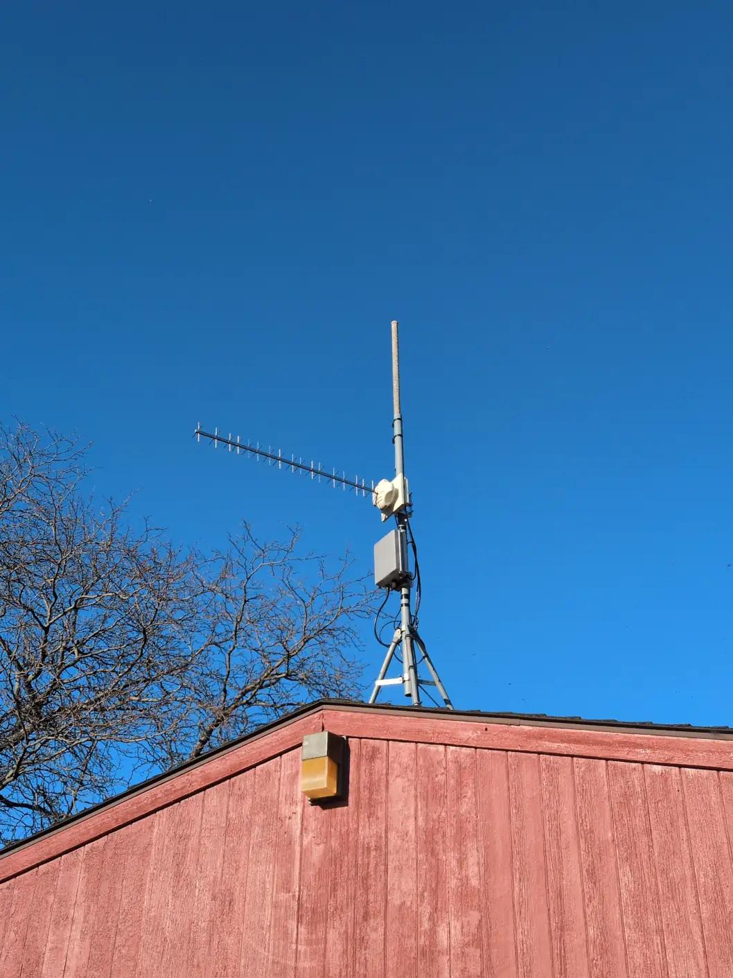 A stand on a roof with a long, horizontal cross-polarized Yagi antenna, a vertical boom, and a box of electronics on the side.