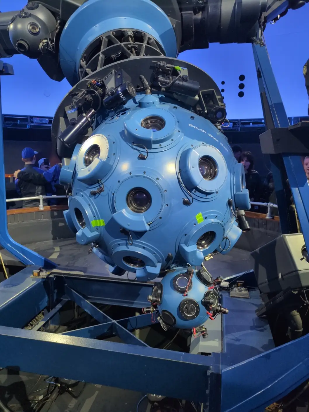 A large blue sphere with raised blue rings on the surface, each surrounding a glass lens. The sphere is mounted on a black metal truss.