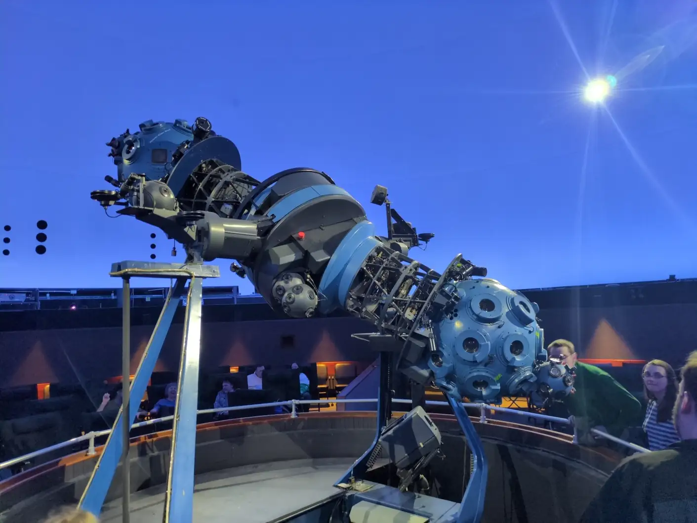 A large cylindrical blue and black metal truss with a pivot passing through the center, held up on a large forked cradle. There is a blue sphere covered in lenses at each end and a number of projectors and lenses mounted on and around the truss.