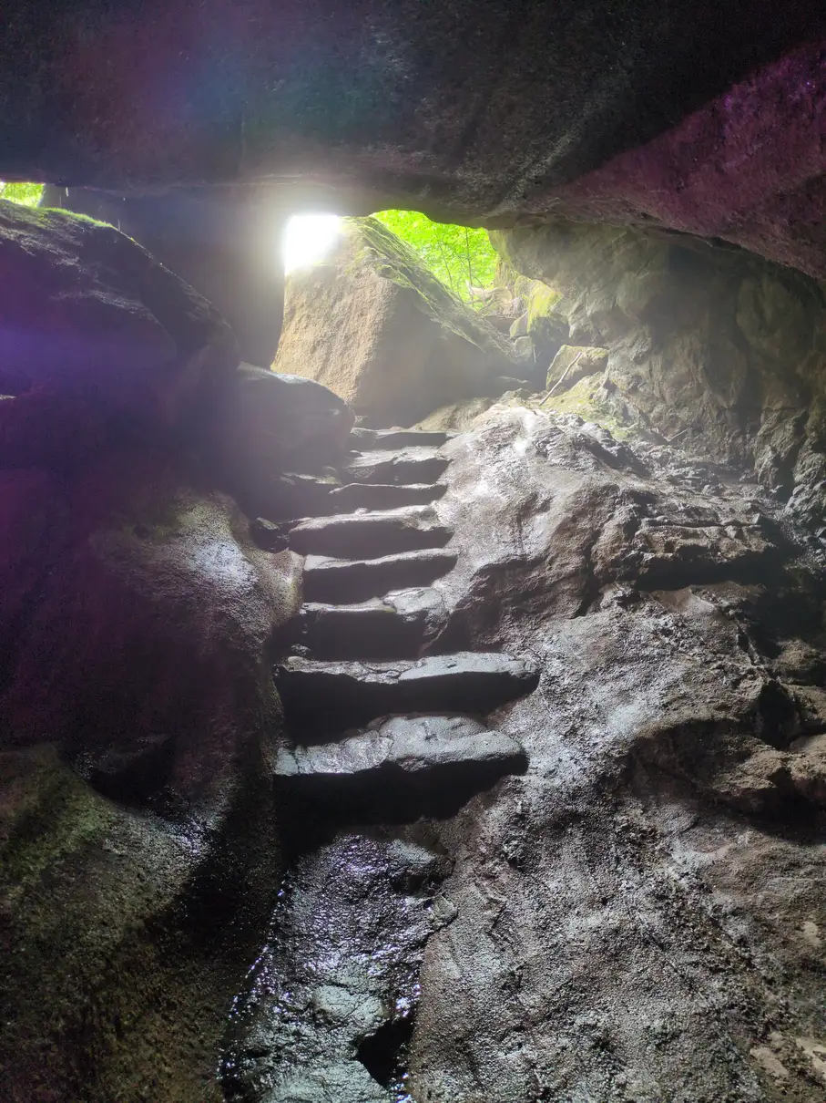 The view out of a rocky cave, with stair steps cut into the ascent and sunlight peeking through the entrance