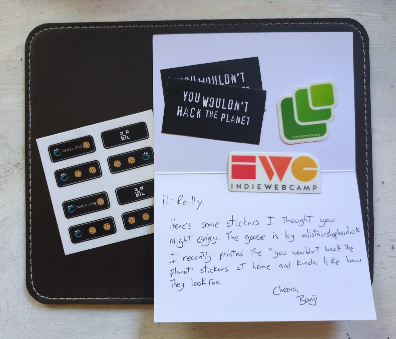 A note with several stickers laid on it. There is a sheet of benji.dog 88x31 banners; a pair of 'you wouldn't hack the planet' stickers; a microformats logo; and an IndieWeb Camp logo.