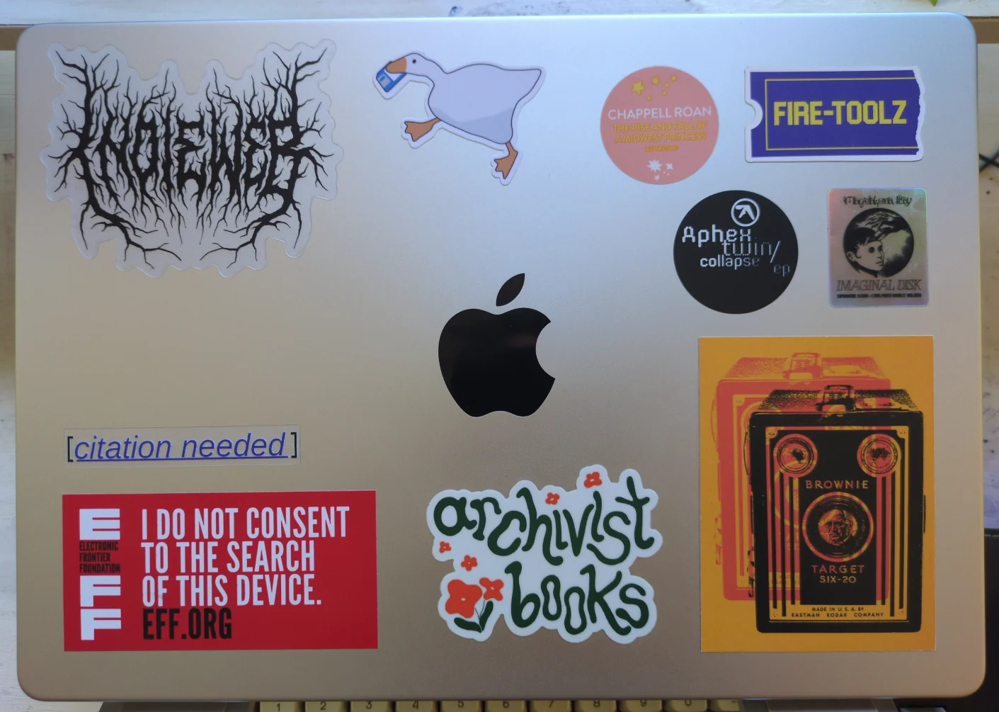 The lid of a silver MacBook, showing numerous stickers. There is a metal band logo-style sticker that says 'IndieWeb'; a goose holding a floppy; stickers from Chappell Roan, Aphex Twin, and Magdalena Bay CD packaging; a Blockbuster ticket that reads 'Fire-Toolz'; a Kodak Brownie Target Six-20 camera; the logo for the local Archivist Books bookstore; an EFF one reading 'I do not consent to the search of this device'; and a Wikipedia 'citation needed' icon from Molly White.