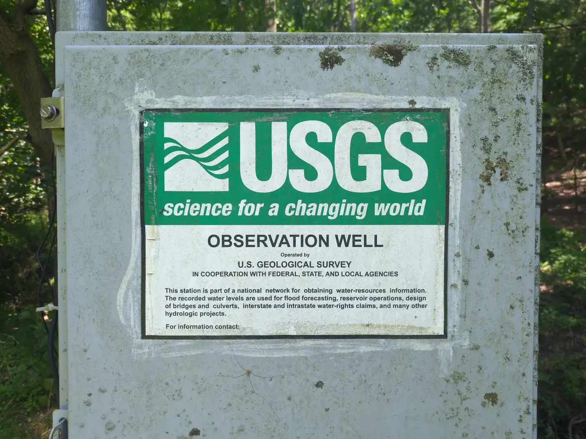 A close up of a rusty gray metal box with a white/green label reading "USGS science for a changing world OBSERVATION WELL Operated by U.S. GEOLOGICAL SURVEY IN COOPERATION WITH FEDERAL, STATE, AND LOCAL AGENCIES This station is part of a national network for obtaining water-resources information. The recorded water levels are used for flood forecasting, reservoir operations, design of bridges and culverts, interstate and intrastate water-rights claims, and many other hydrologic projects. For information contact:"